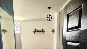 Ein gemütliches Schlafzimmer mit Bett, Nachttisch und kleinem Stuhl. Ein Fenster mit dekorativem Sichtschutz und ein Regal mit Pflanzen sind ebenfalls vorhanden.