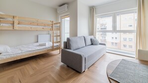Una habitación con una cama litera, un sofá, una ventana con vista a edificios y un ventilador de techo.