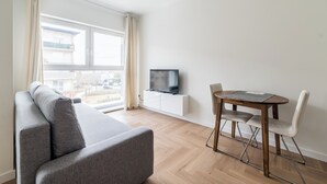 Una sala de estar moderna con un sofá gris, una mesa de comedor de madera con sillas, un televisor sobre un mueble blanco y un ventanal con cortinas.