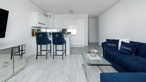 Un salon moderne avec un canapé bleu, deux tabourets de bar, une table basse en verre et une cuisine en arrière-plan.
