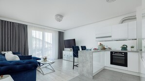 Un salon moderne avec un canapé bleu, une table basse en verre et une cuisine dotée de meubles blancs et d’un four noir.