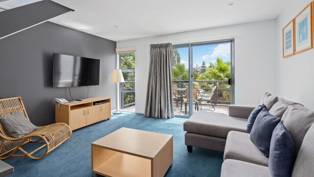 A modern living room with a flat-screen TV, a sofa, a coffee table, and a wicker chair. - 2 Bedroom Open plan,combined living & dining areas with fully self contained kitchen & laundry. (Batehaven)