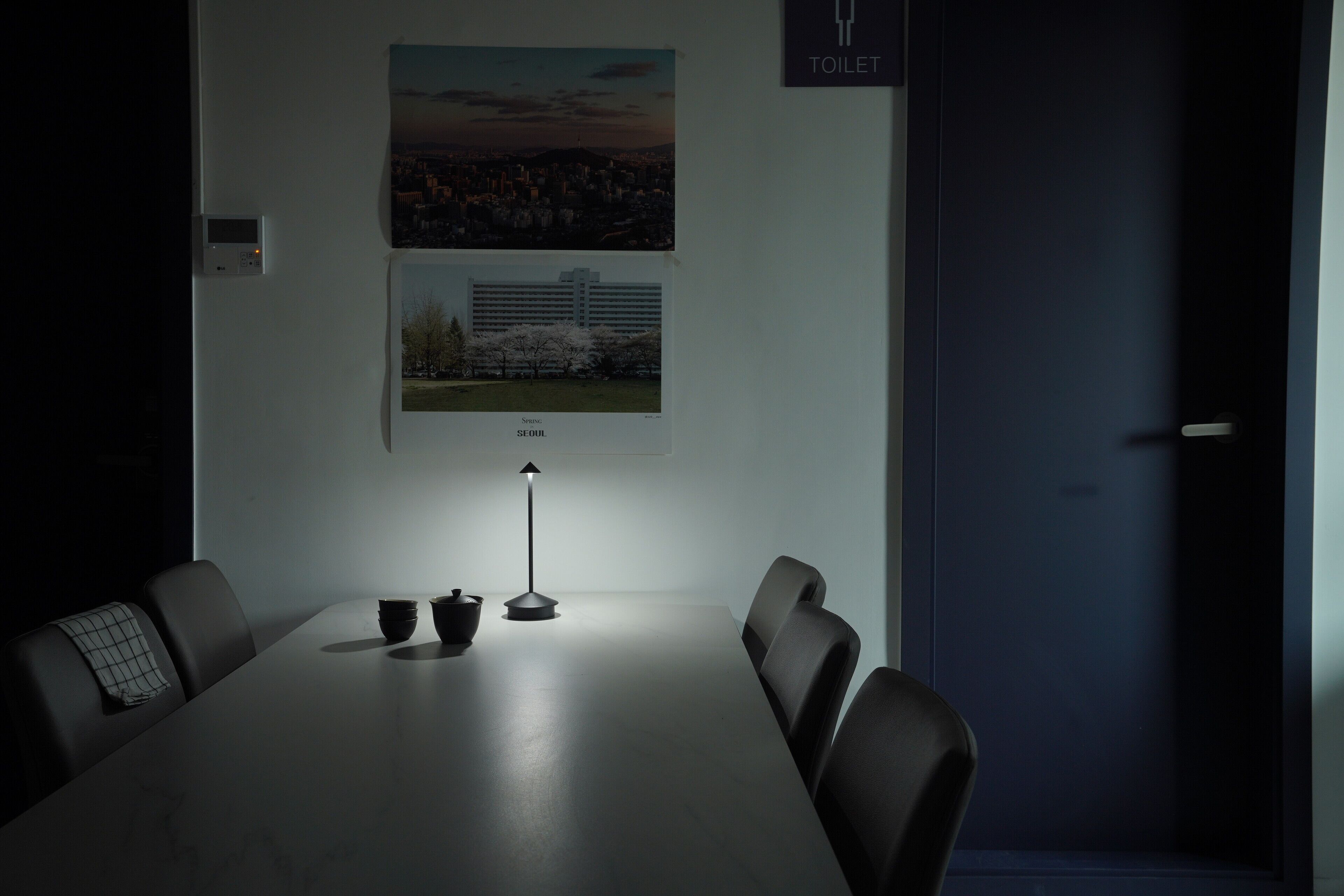 A conference room with a long table, four chairs, a small table lamp, two cups, and two framed pictures on the wall.