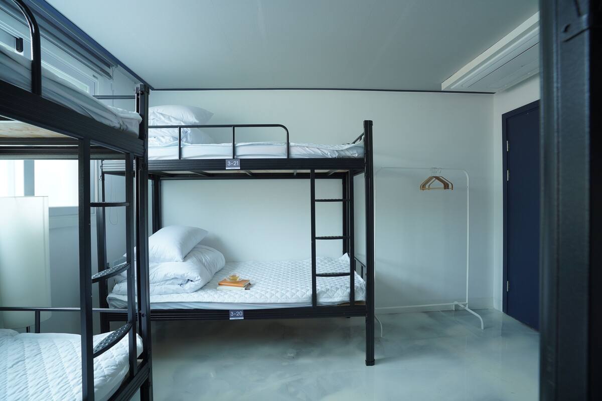 Een slaapkamer met een stapelbed, een witte matras, een klein tafeltje met een boek en een deur.
