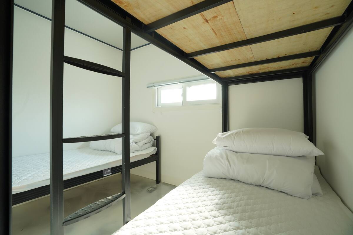 Een slaapkamer met stapelbed, een raam, metalen balken en een wit matras.