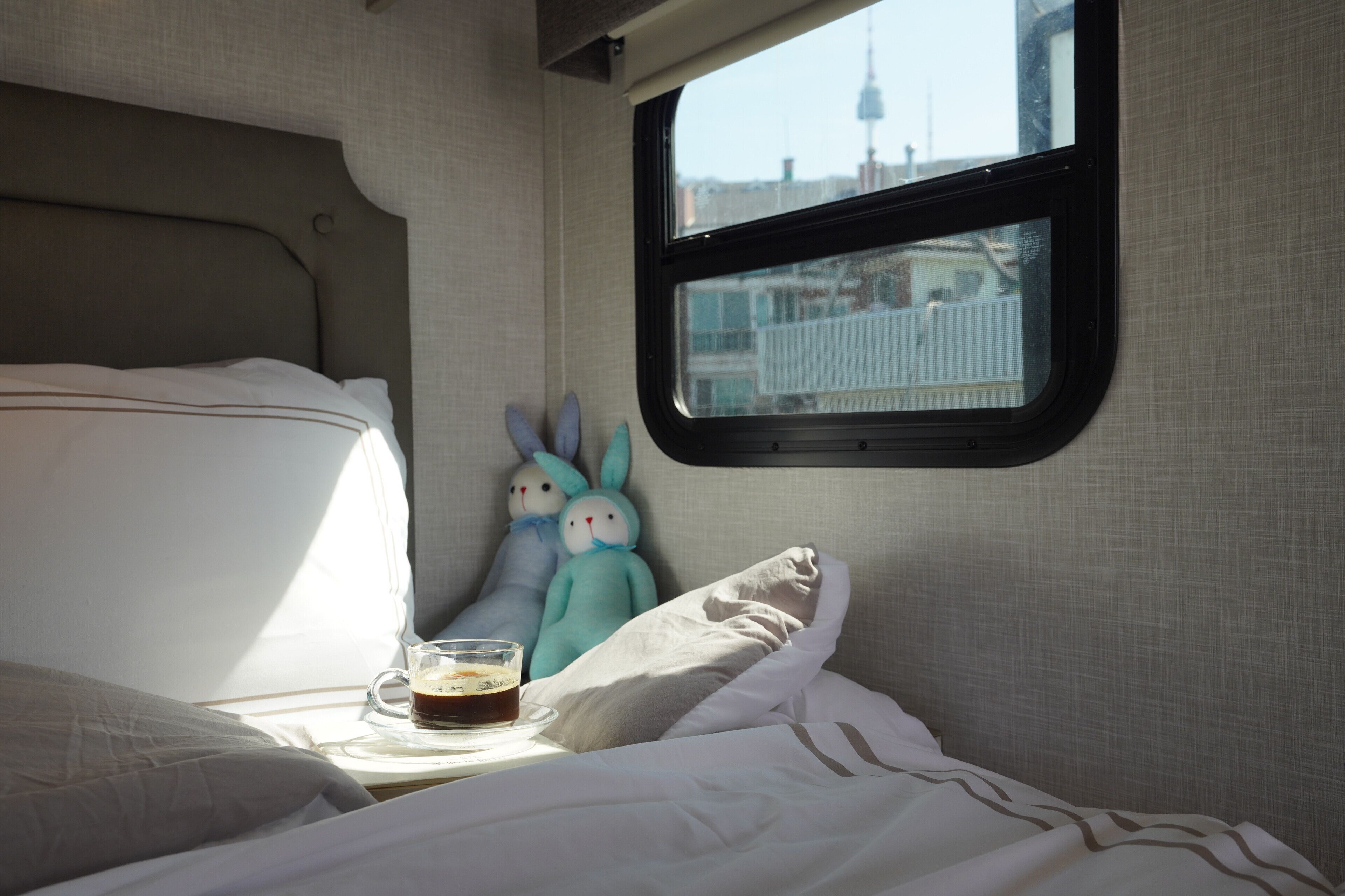 A bed with pillows, a glass of coffee, and two plush toys on a bedside table.