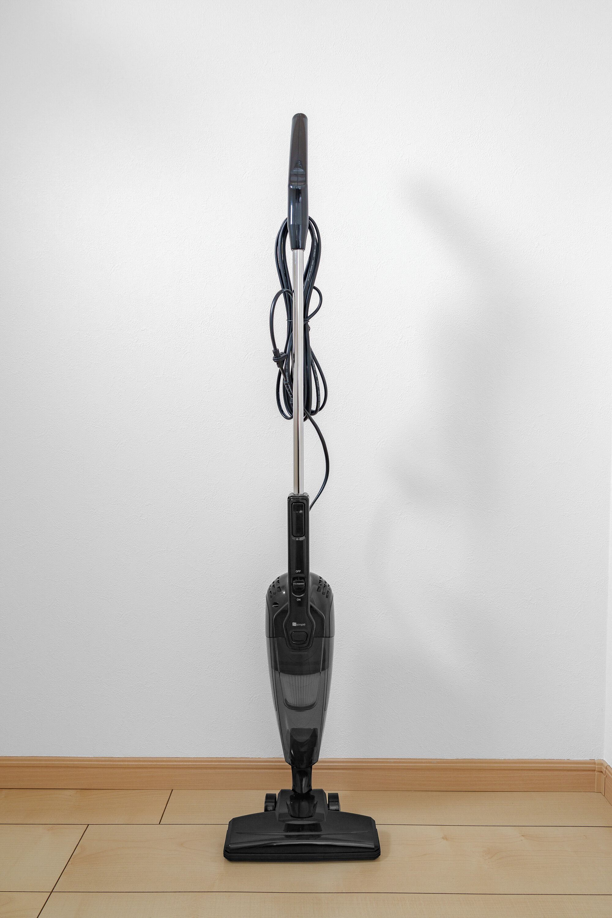 A standing vacuum cleaner with a cord, placed on a wooden floor against a white wall.