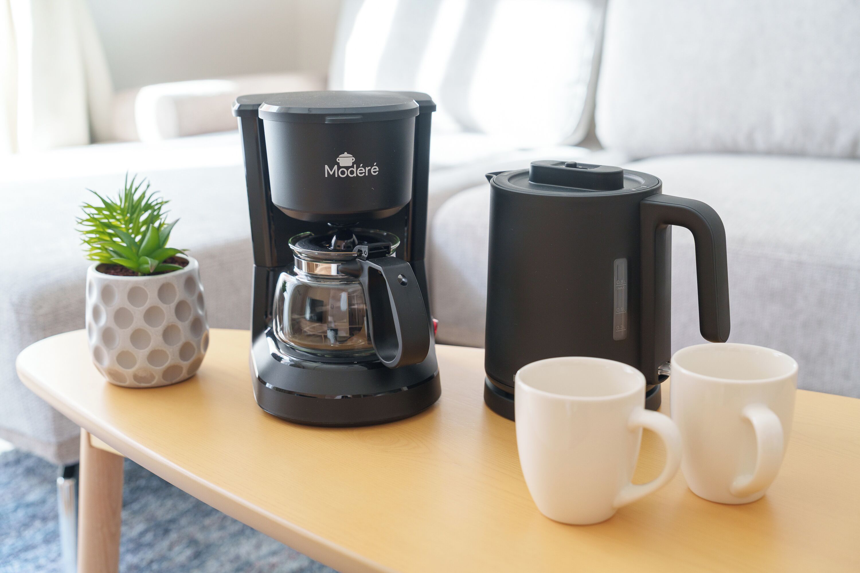 Eine Kaffeemaschine, zwei weiße Tassen und ein kleiner Topfpflanze auf einer Holztisch.