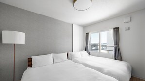 Habitación de hotel con dos camas, un ventanal con cortinas, una lámpara de pie y vistas a la ciudad.
