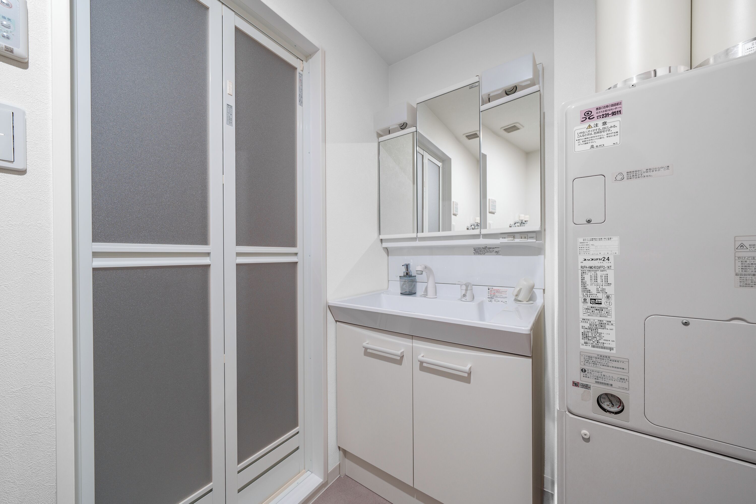 A small, white bathroom with a frosted glass door, a white sink with a mirror above it, and a white refrigerator with a water filter.
