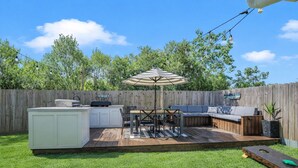 Un giardino sul retro con un patio in legno, un barbecue, un tavolo da pranzo con sedie, un ombrellone grande e una zona relax integrata.