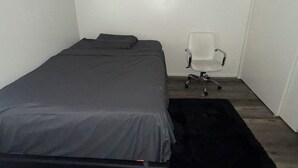 Una habitación pequeña con una cama individual, una colcha gris, una silla de oficina blanca y un tapete negro.