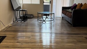Amplia sala de estar con piso de madera, una mesa redonda de vidrio, un sofá y un televisor sobre un mueble.