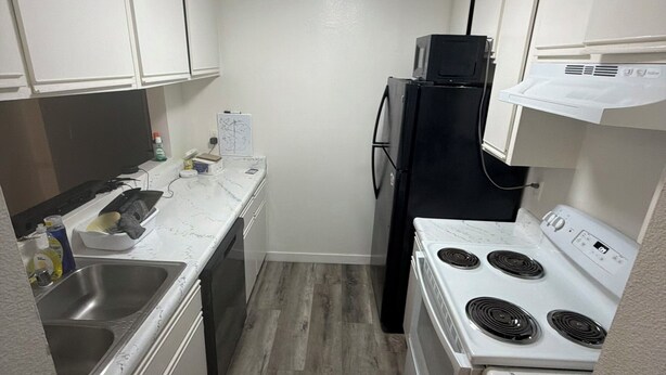 Una cocina compacta con armarios blancos, un refrigerador negro, una estufa blanca y un lavaplatos de acero inoxidable.