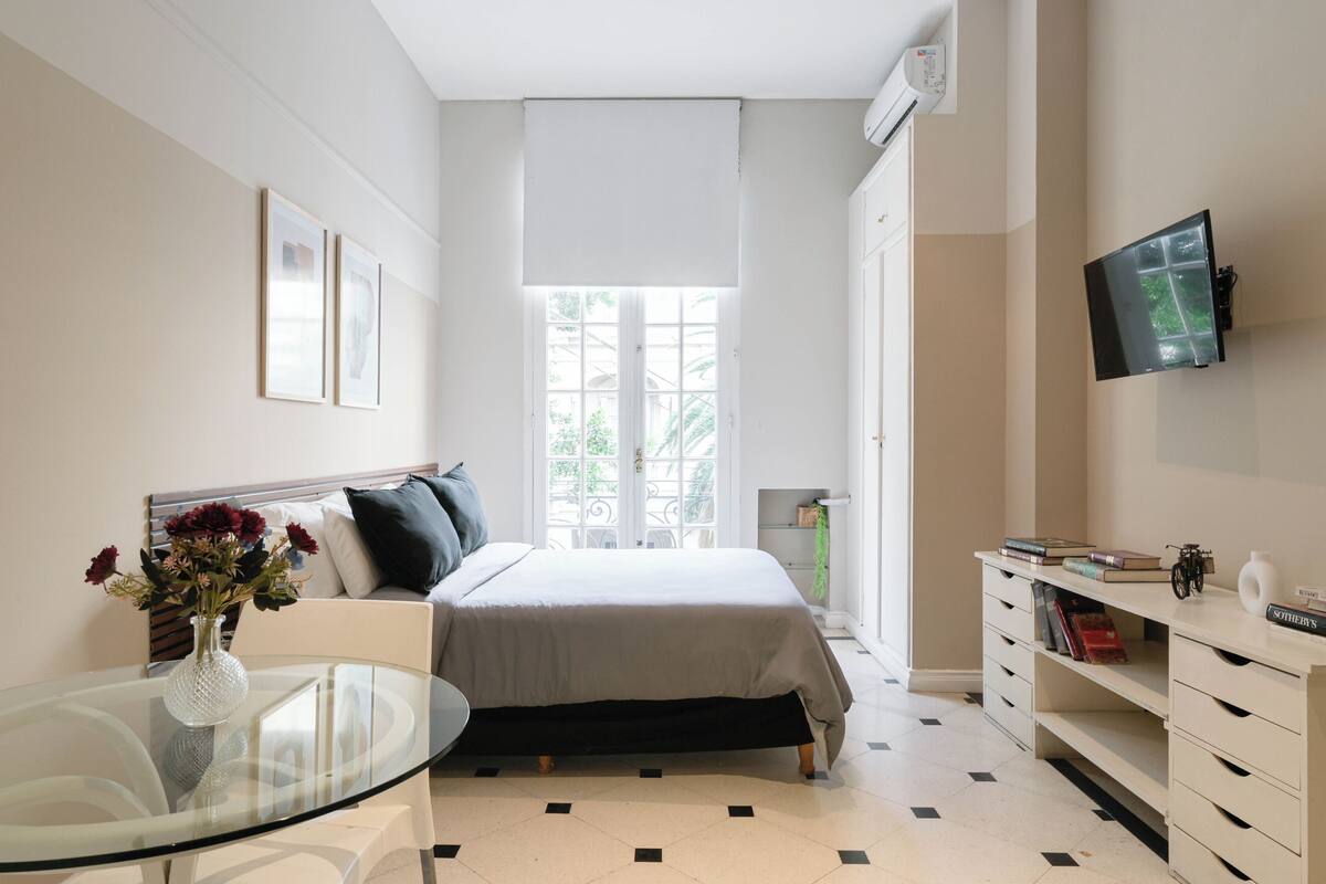 Ein Schlafzimmer mit einem Bett, einem Nachttisch mit Blumenvase, einem an der Wand befestigten Fernseher und einem Fenster mit Blick auf Grünflächen.