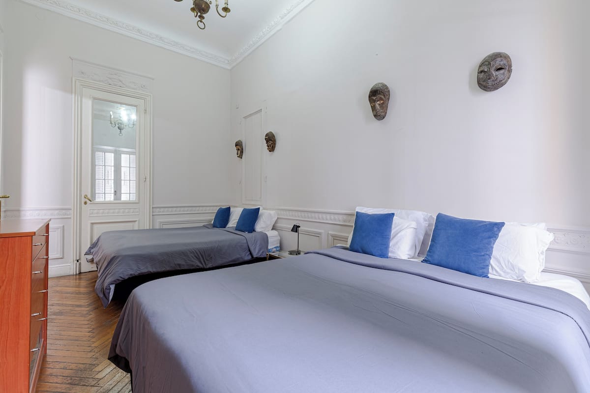 Habitación de hotel con dos camas, cómoda de madera, candelabro y máscaras decorativas en la pared.