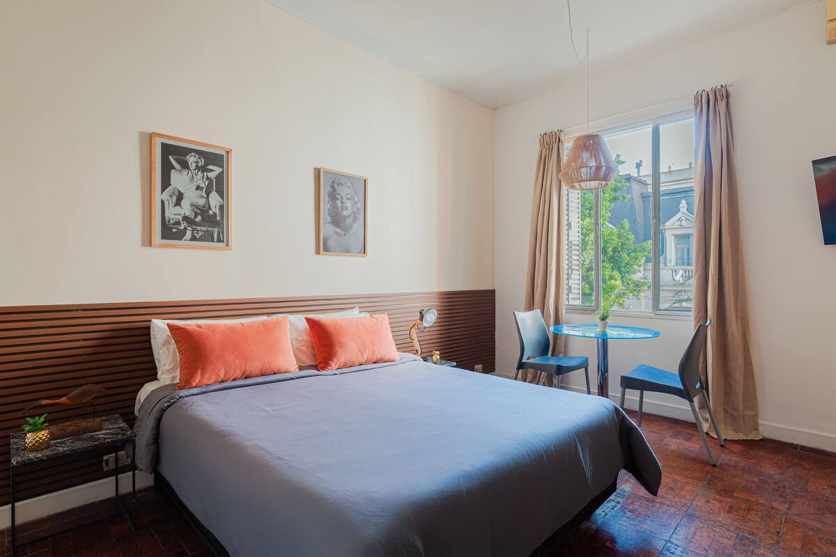 Ein Hotelzimmer mit einem Bett, zwei gerahmten Bildern, einem Fenster mit Vorhängen, einem kleinen Tisch mit einem Stuhl und Blick auf Gebäude.