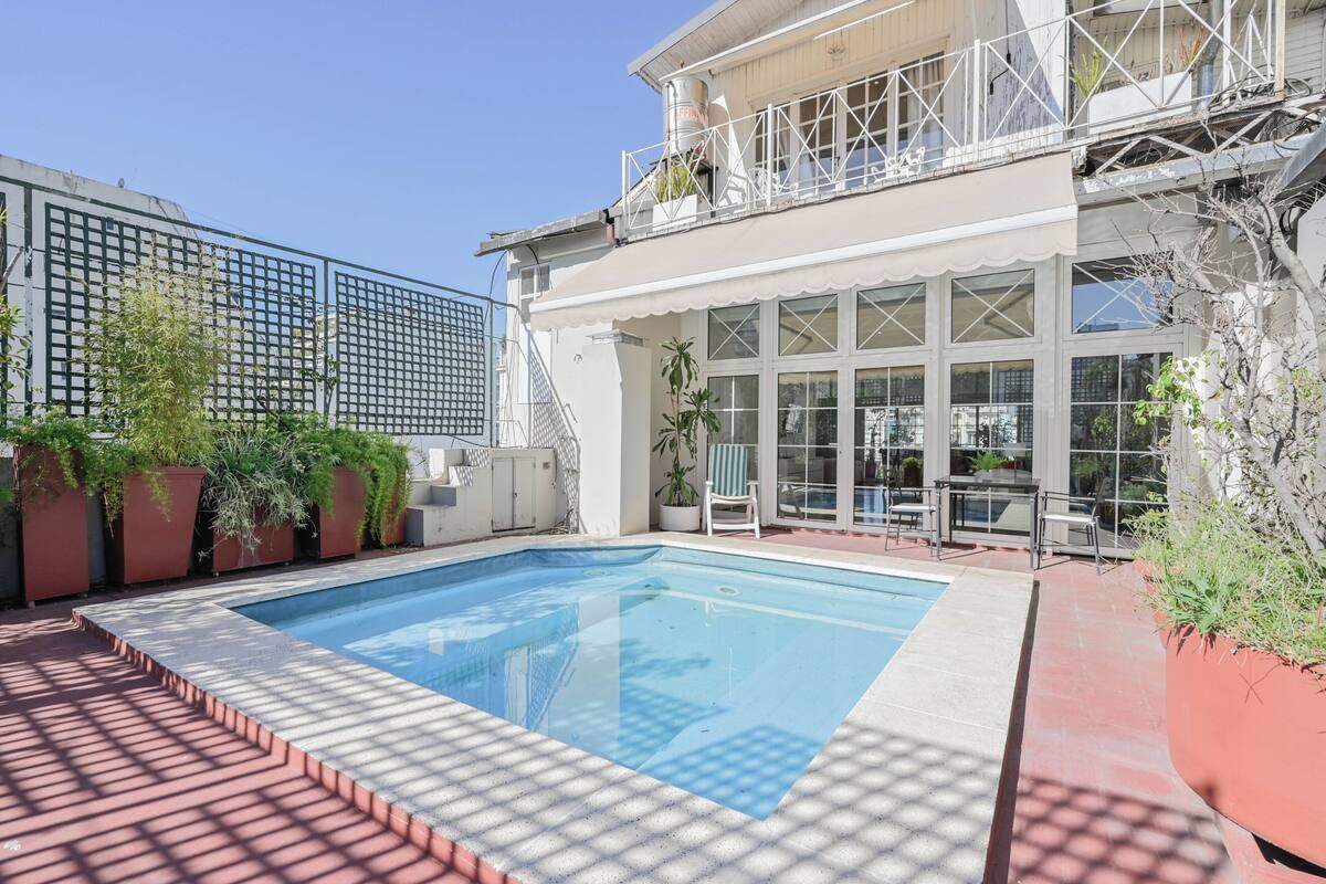 Ein Schwimmbad mit gefliestem Deck, ein Balkon mit Sitzgelegenheiten und ein großes Zimmer mit Fenstern.