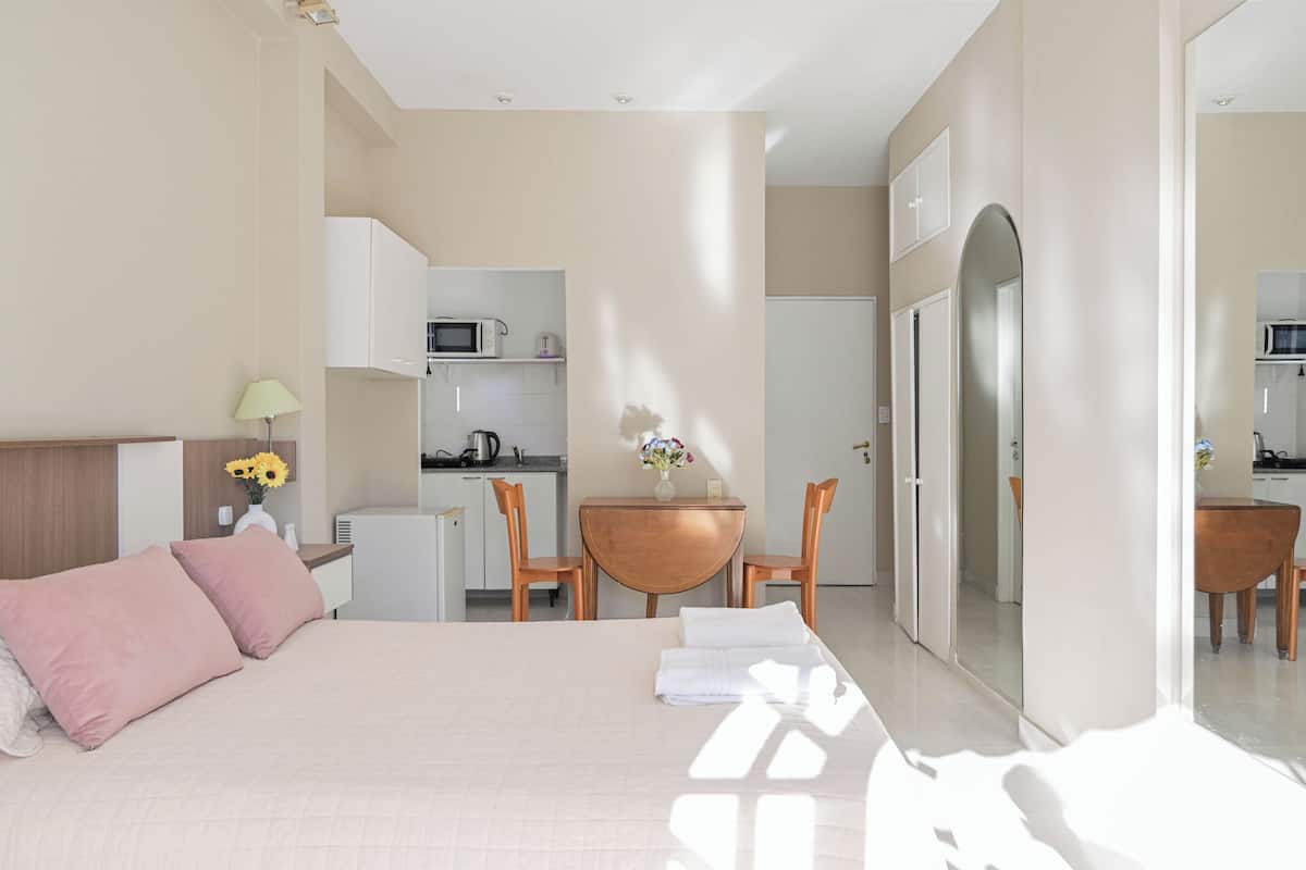 Un dormitorio con cama, escritorio y kitchenette al fondo.