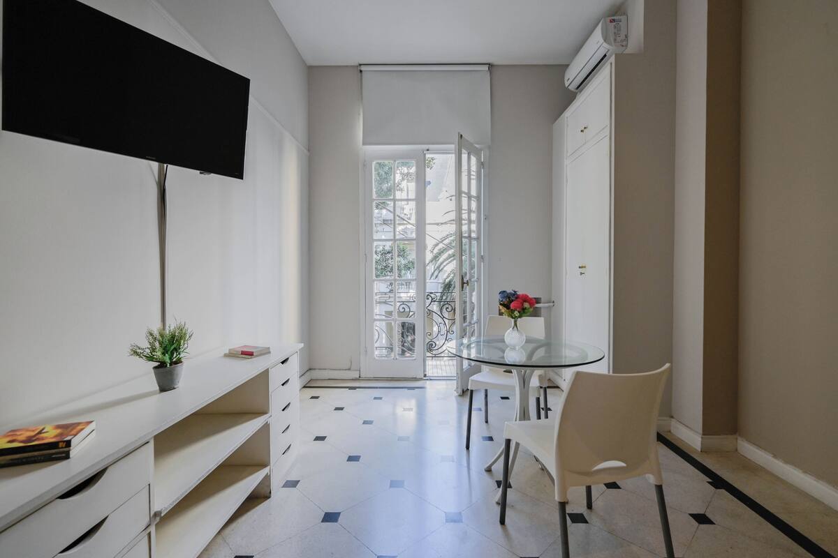 Ein modernes Interieur mit einem an der Wand montierten Flachbildfernseher, einem Glastisch mit Stühlen, einem Topf mit Pflanze und einer zur Außenseite führenden Tür.