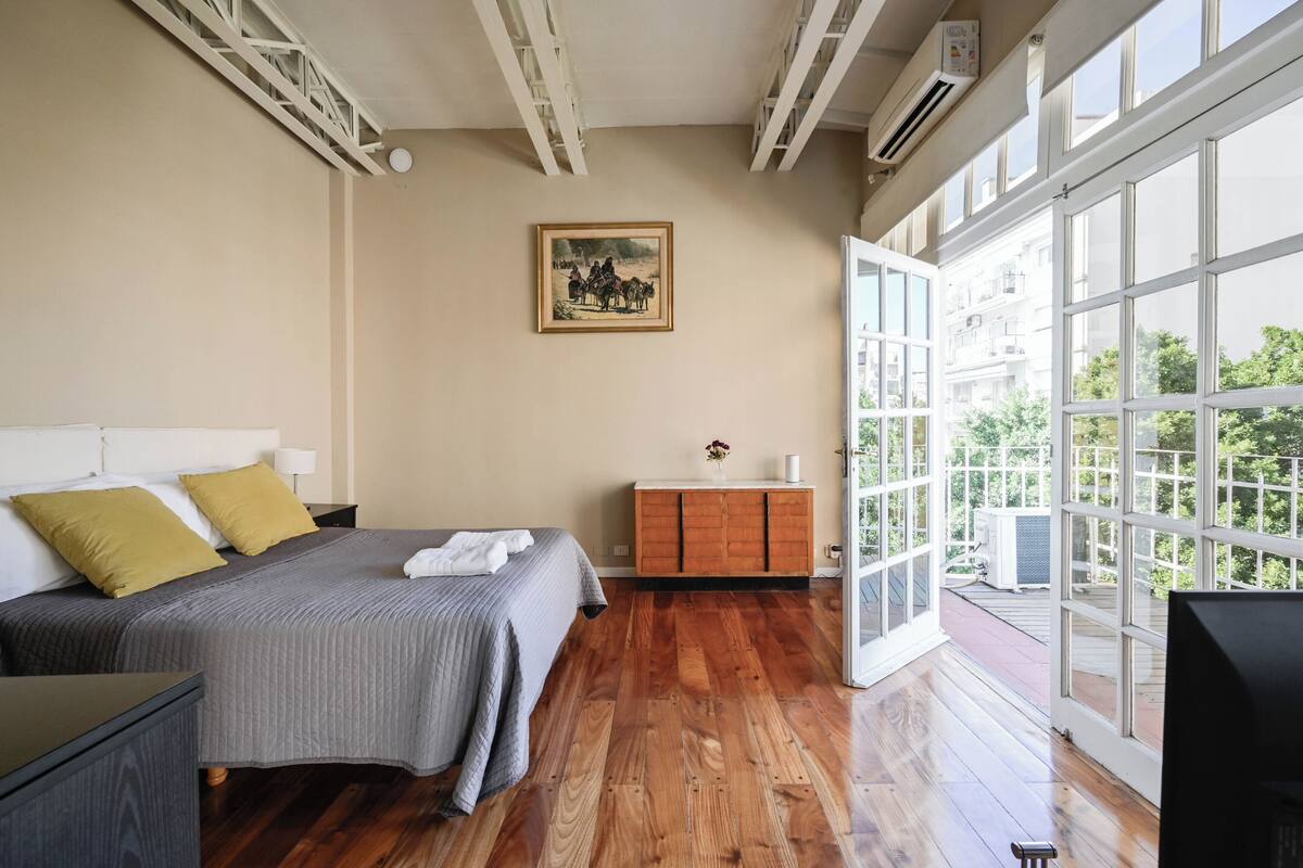 Ein Schlafzimmer mit Bett, Nachttisch, Kommode, einem Gemälde und einem großen Fenster.