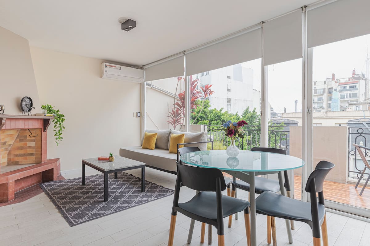 Una sala de estar moderna con una mesa de vidrio, sillas negras, una chimenea y vistas de la ciudad a través de grandes ventanales.