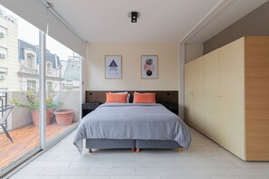 Une chambre à coucher avec un lit, deux tableaux encadrés au mur, une armoire en bois et un balcon agrémenté de plantes en pot.