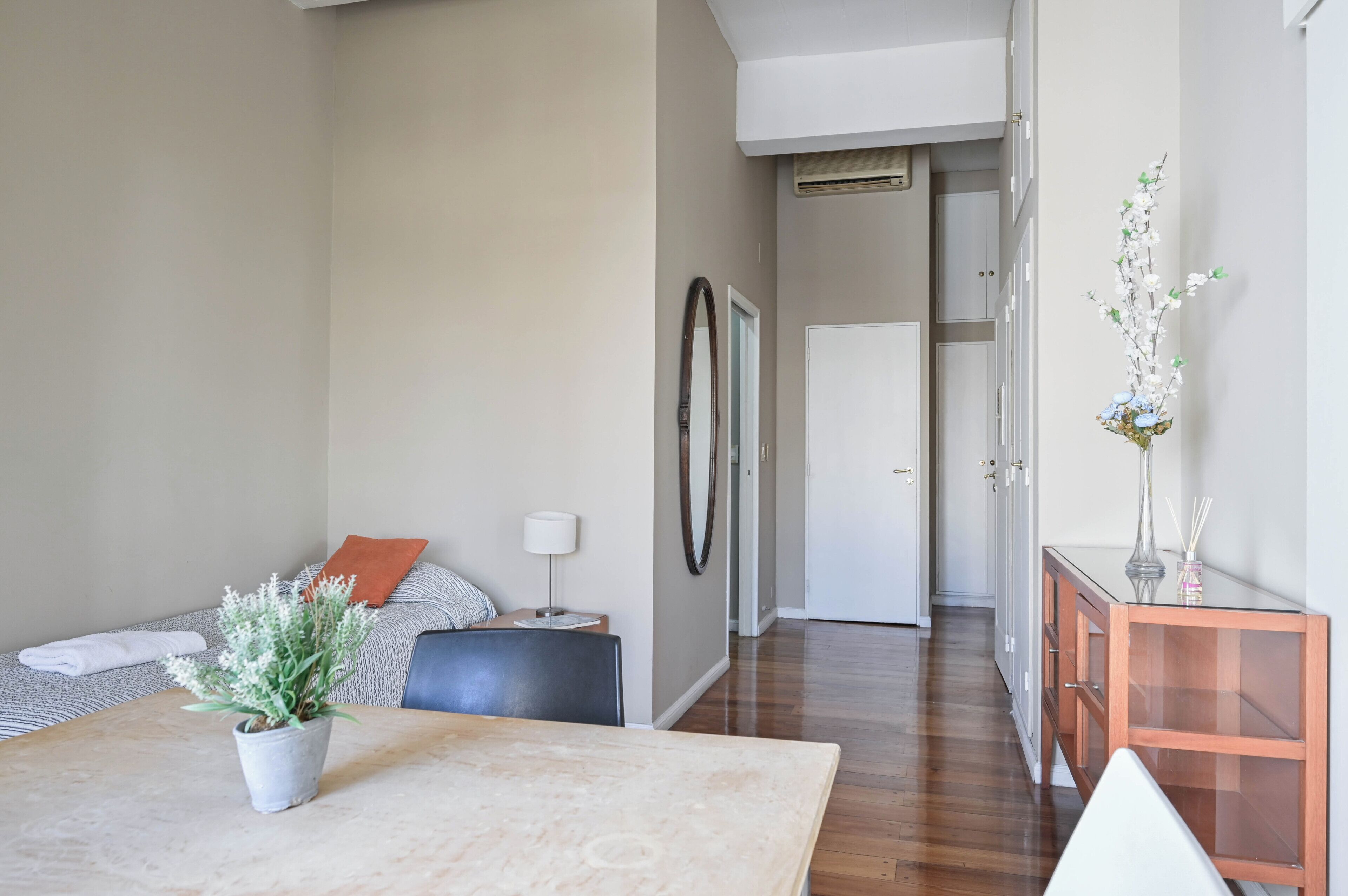 Een moderne hotelkamer met een houten vloer, een eettafel, een bank, een klein bijzettafeltje met een lamp, en een gang naar een deur.