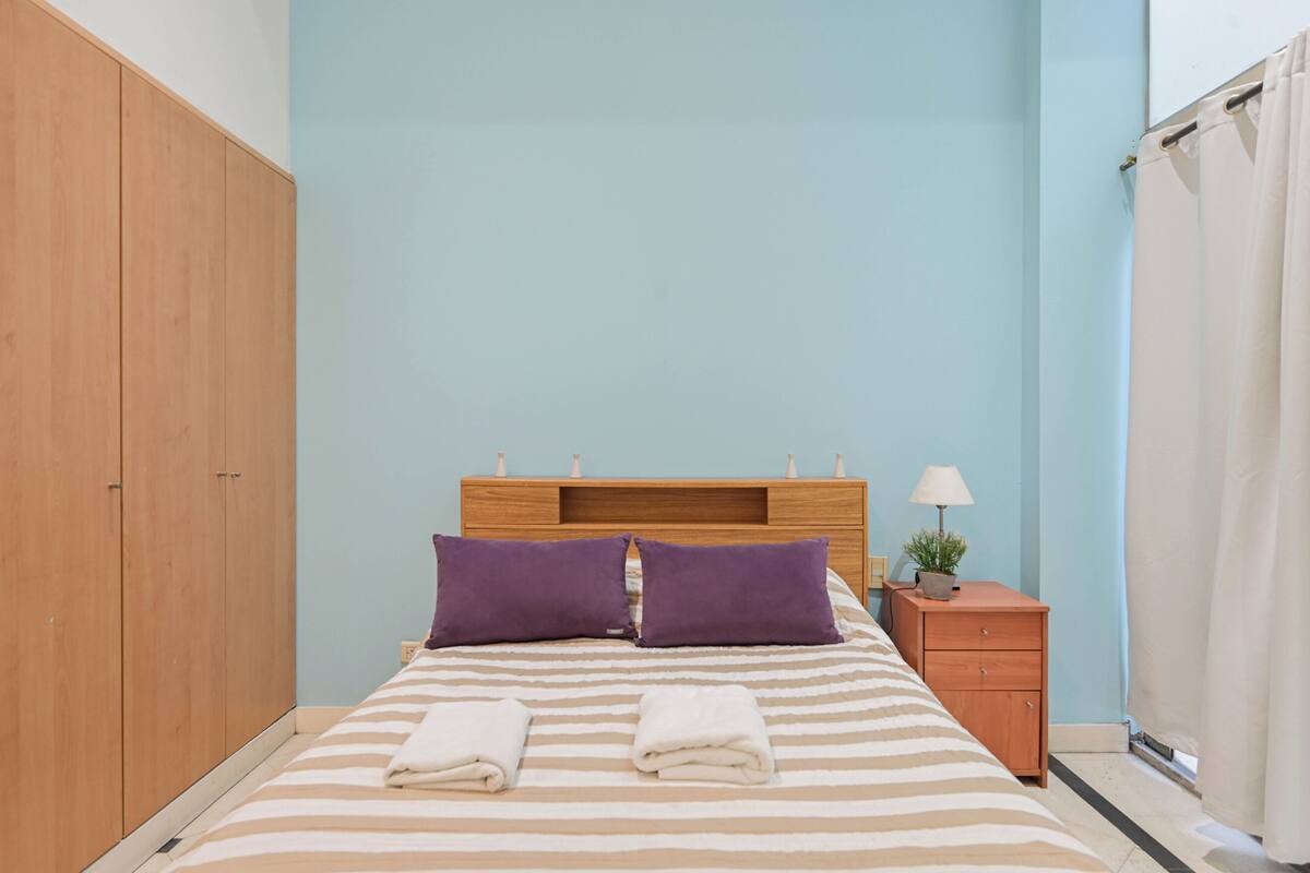 Ein Schlafzimmer mit einem Bett, zwei Kissen, einem Nachttisch mit Lampe, einer kleinen Pflanze und einem Holzschrank.