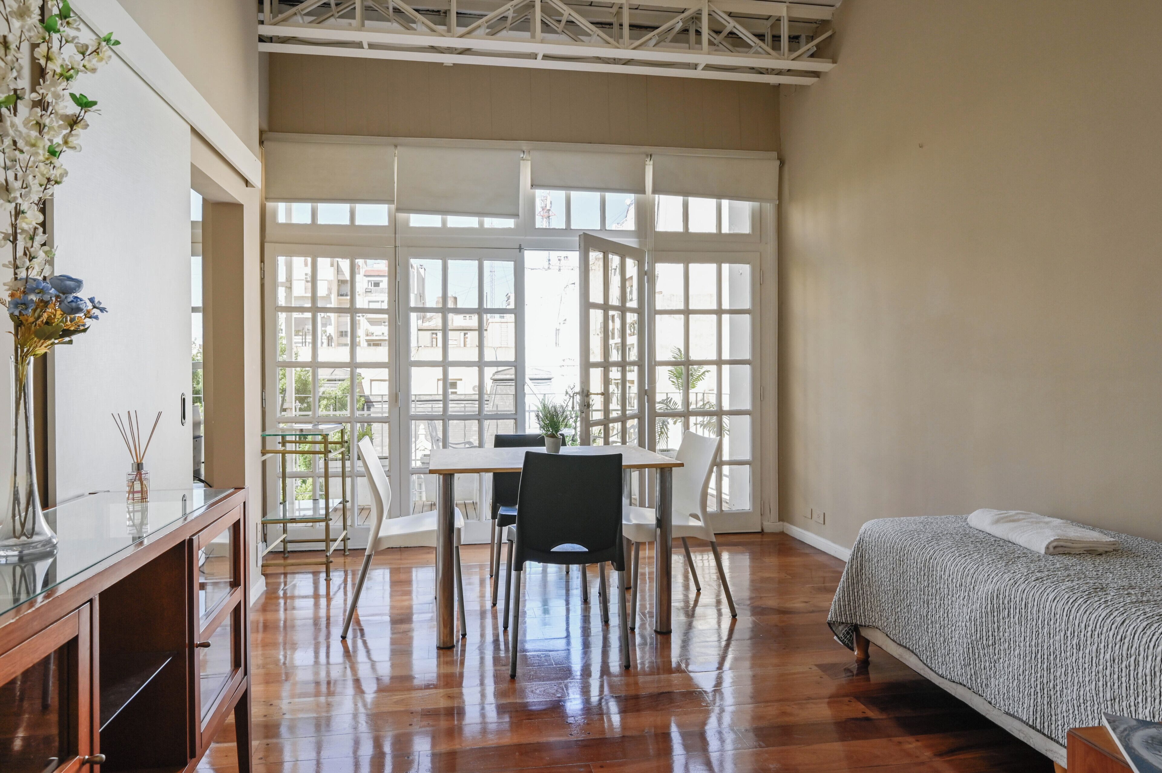 A room with a bed, a dining table with chairs, a vase with flowers, and large windows.