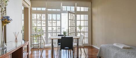 A room with a bed, a dining table with chairs, a vase with flowers, and large windows.