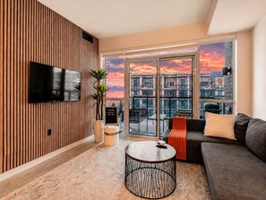 A modern living room with a TV, a round coffee table, a sofa, and a view of a cityscape through large windows. - Stunning 1BR w Pool ~ Free Parking & Self Check-In (Innisfil)