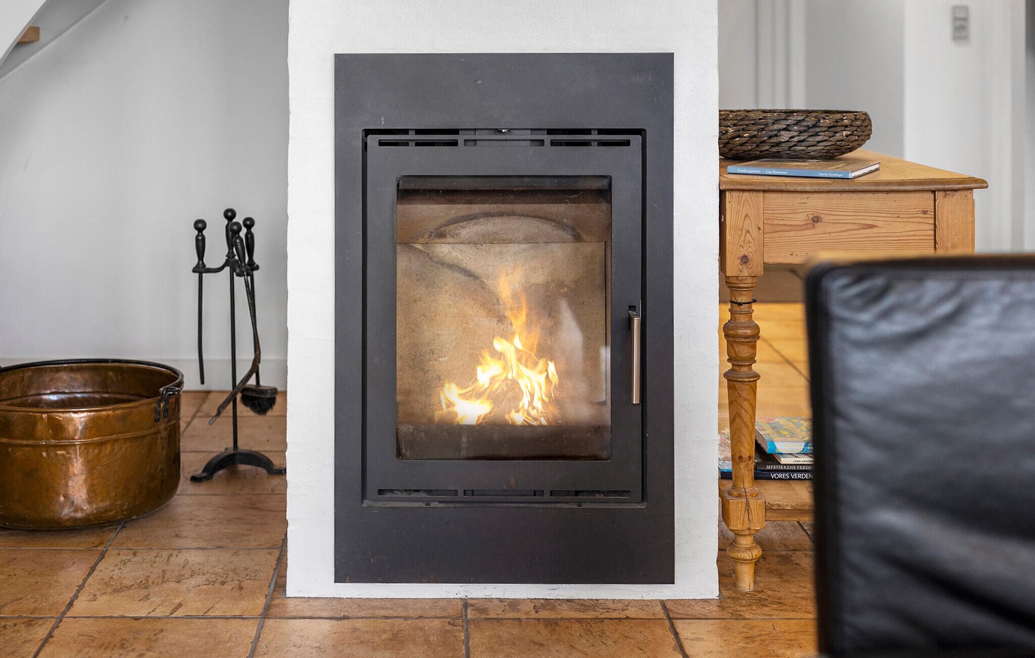 Una chimenea moderna con puerta de cristal, un mueble de madera con una cesta tejida, y un balde de metal sobre el suelo.