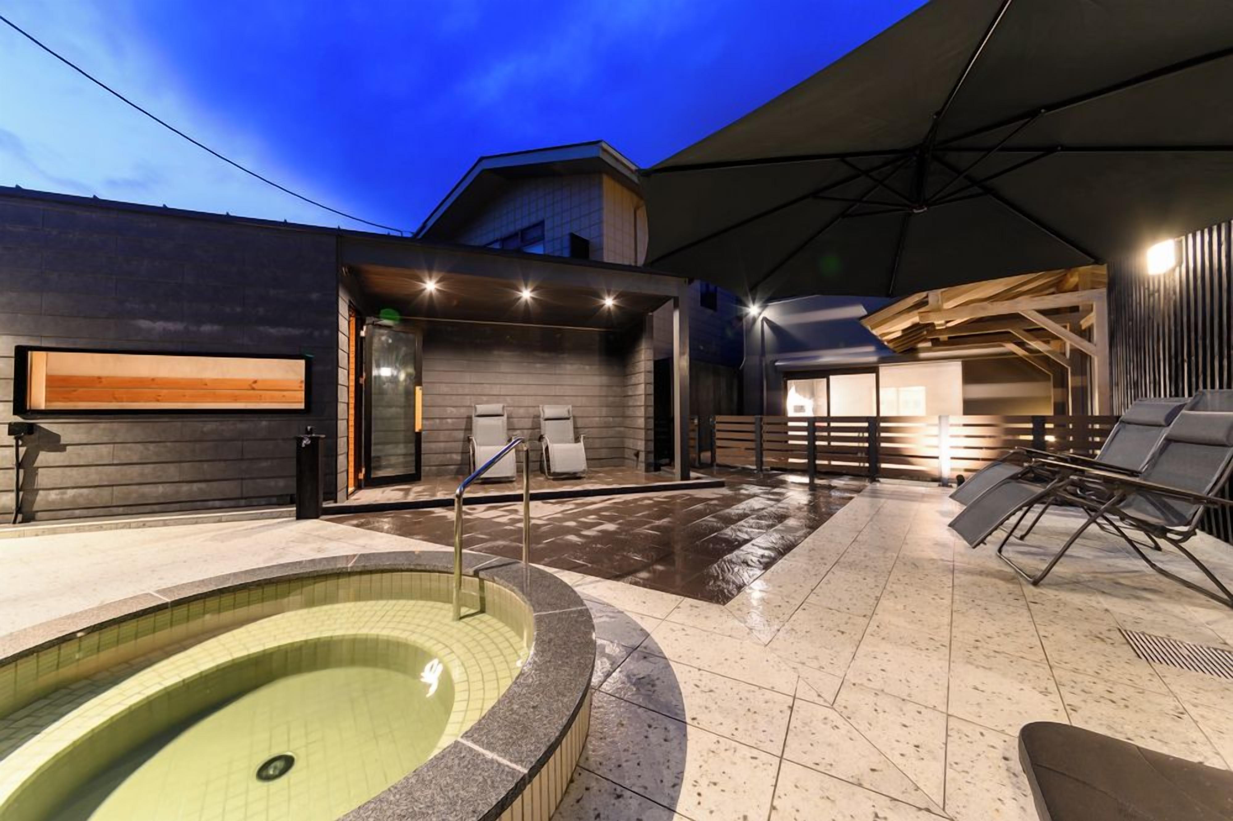 Un área de piscina moderna al aire libre con jacuzzi, sillas de descanso y una zona de estar cubierta.