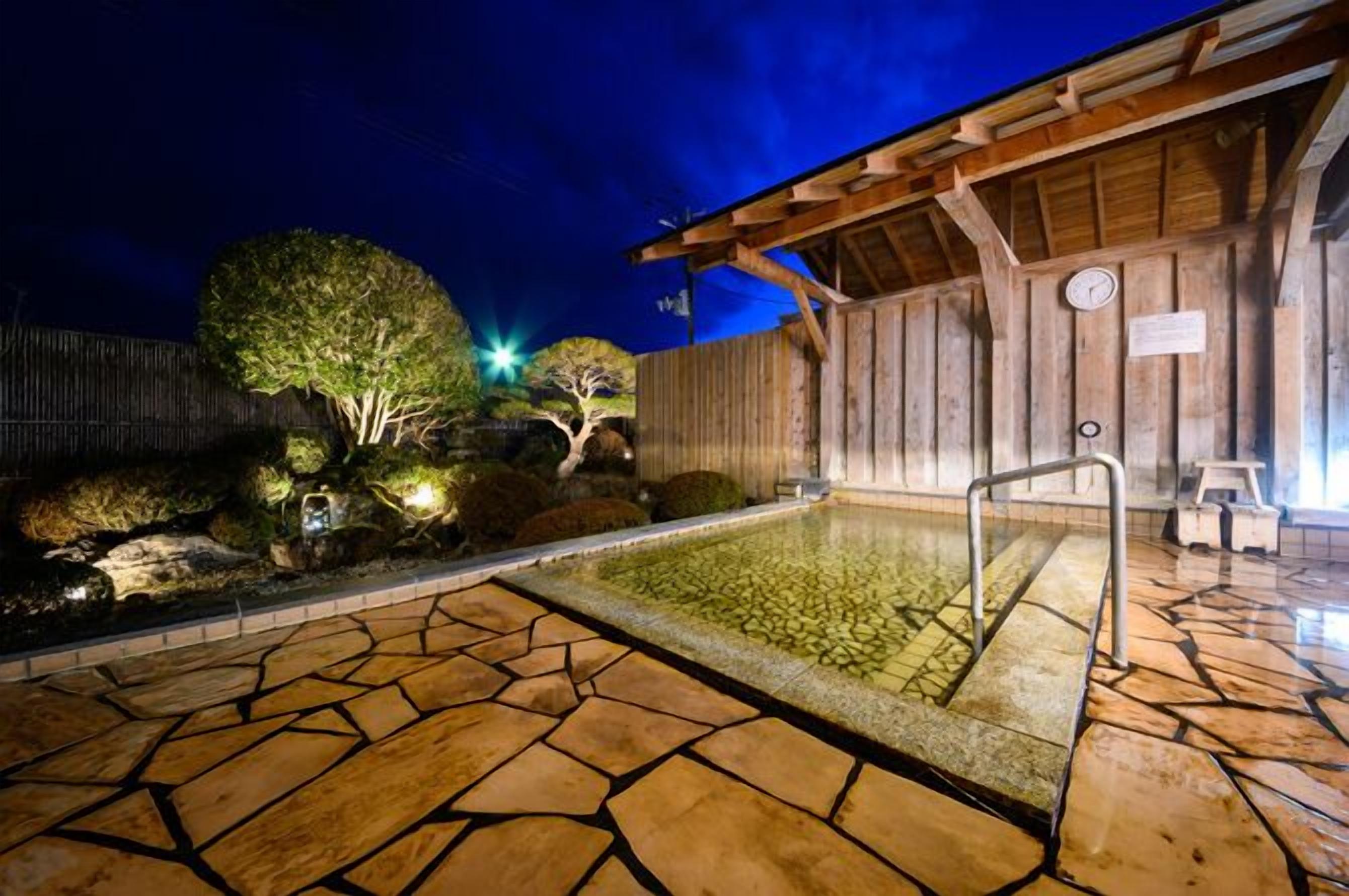 Área de piscina ao ar livre com piso de pedra, estrutura de madeira, um relógio e um pequeno espaço com assentos.
