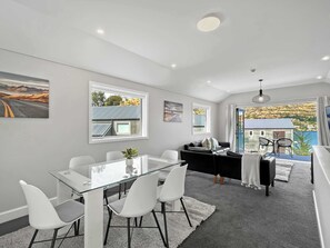 Een moderne woonkamer met een glazen eettafel, witte stoelen, een zwarte bank en uitzicht op een kustlandschap.