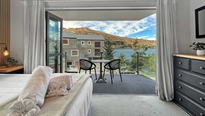 Un dormitorio con cama, cómoda, una mesa con sillas y vistas a un edificio y montañas a través de una puerta de cristal.