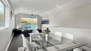 Una habitación moderna con una mesa de vidrio, sillas blancas, un sofá, un televisor y vistas a un paisaje montañoso.