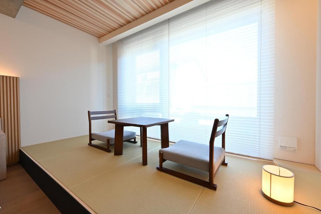 Una habitación con tatamis, una mesa baja, dos sillas, una lámpara y una ventana con persianas.