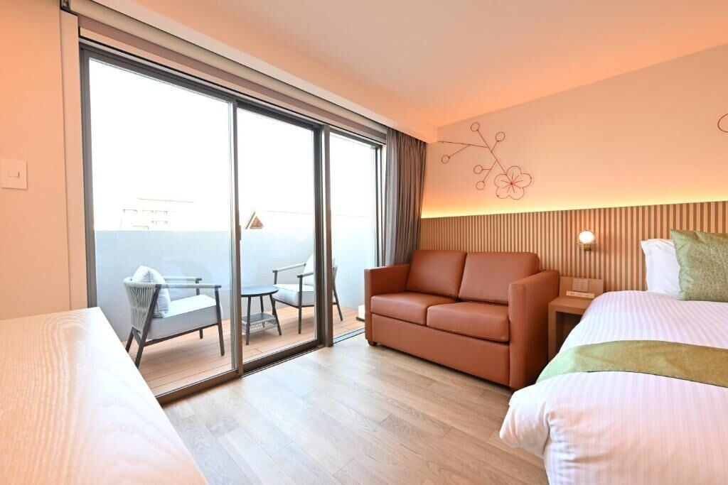 A hotel room with a bed, a sofa, a small table, and a chair. There is a sliding glass door leading to an outdoor area.