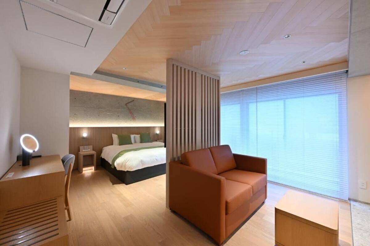 A modern hotel room with a bed, a sofa, a desk with a mirror, and a large window.