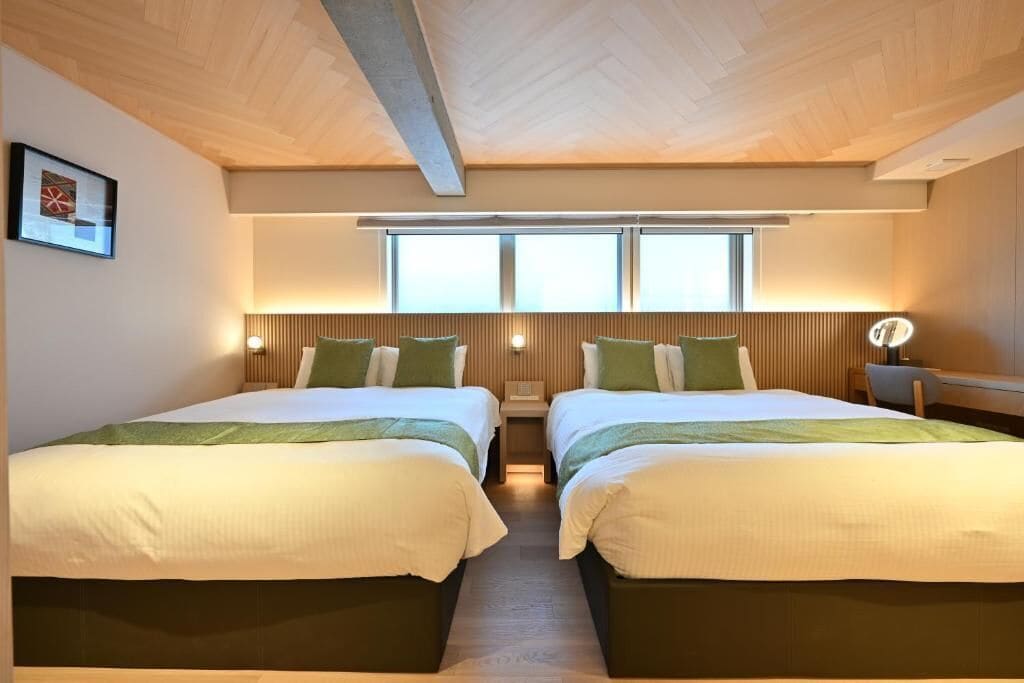 A hotel room with two beds, wooden ceiling, large windows, and a nightstand with a lamp.