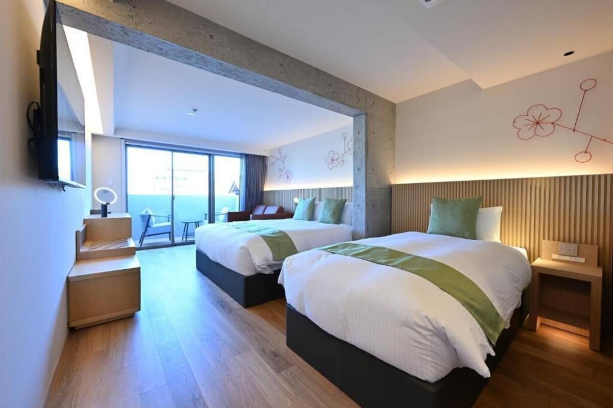 A modern hotel room with two beds, a desk, and a view of the outdoors.