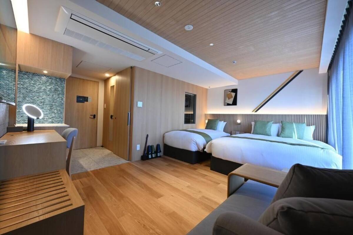 A modern hotel room with a wooden ceiling, a desk with a mirror, a bed with a green headboard, and a large window.