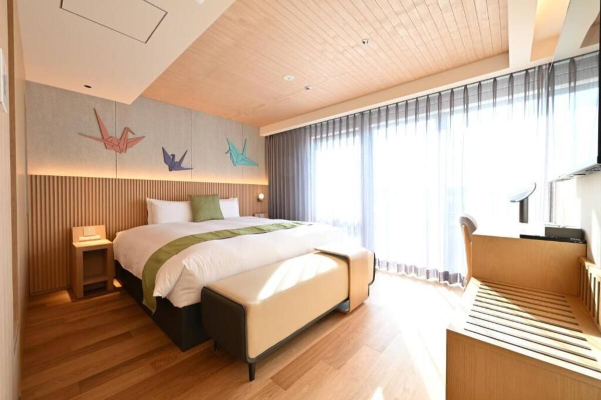 A modern hotel room with a wooden floor, a large bed with a green throw, a bedside table, a chair, and a large window with curtains.