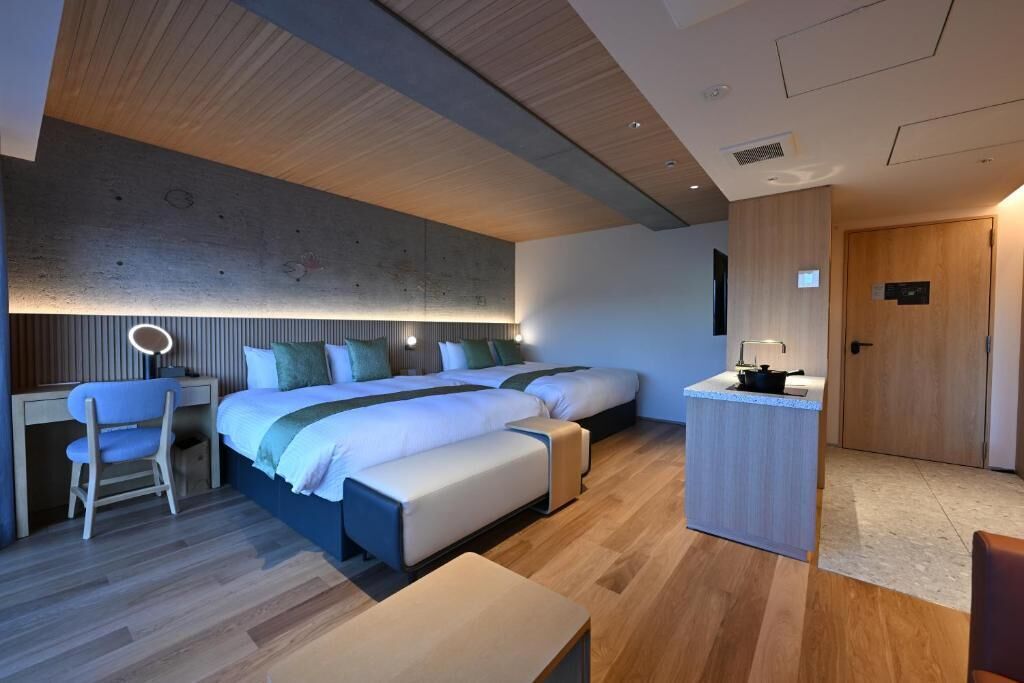 A modern hotel room with a large bed, a desk, a chair, a small kitchenette, and a wooden ceiling.