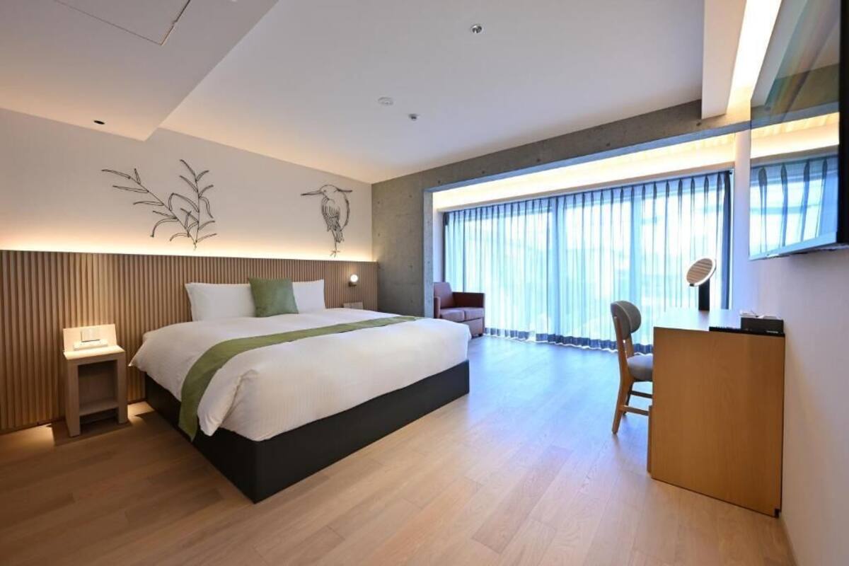 A modern hotel room with a large bed, a desk, and a chair, a large window with curtains, and a wall-mounted artwork.
