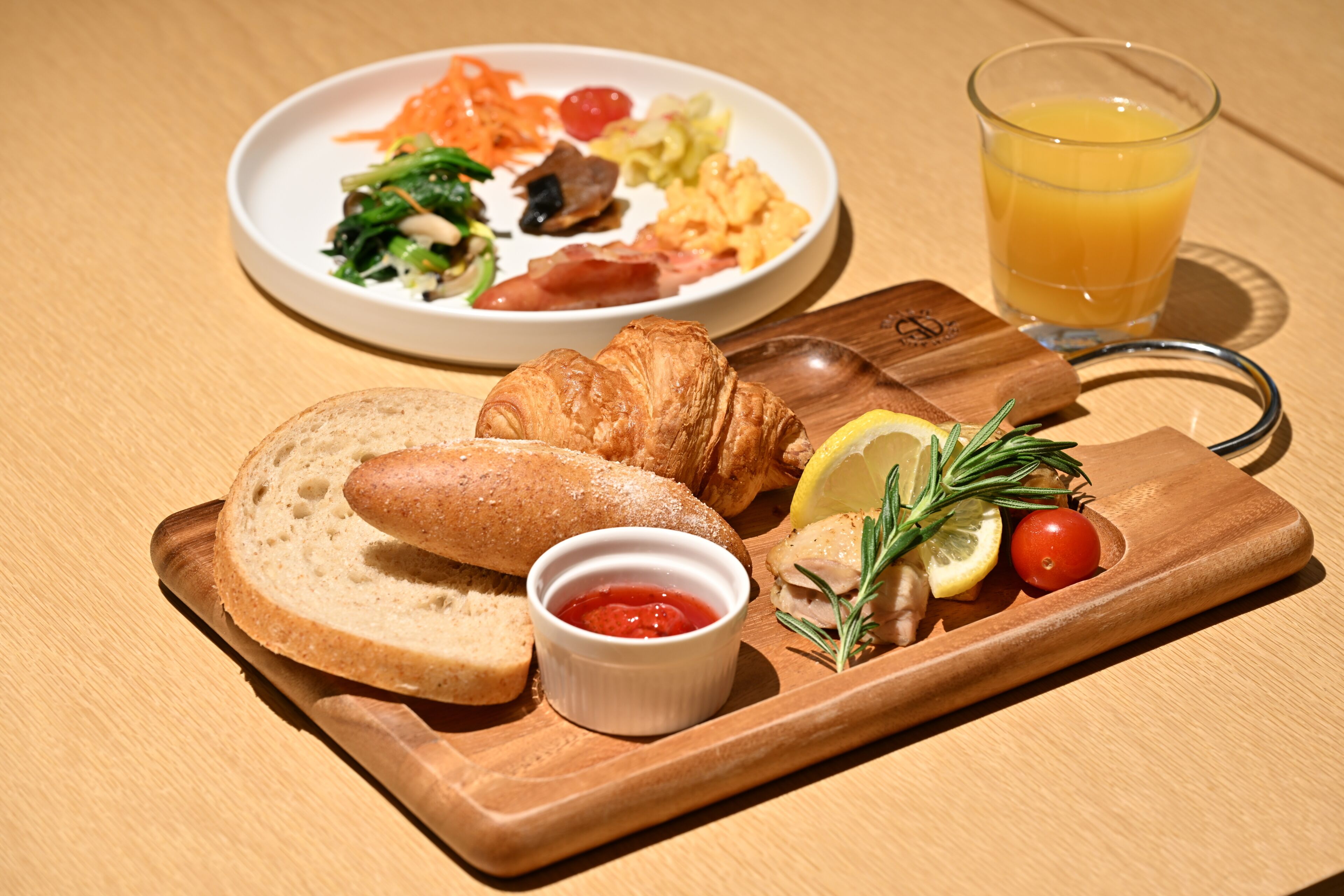 Uma bandeja de café da manhã com pão, croissant, vegetais e um copo de suco de laranja.