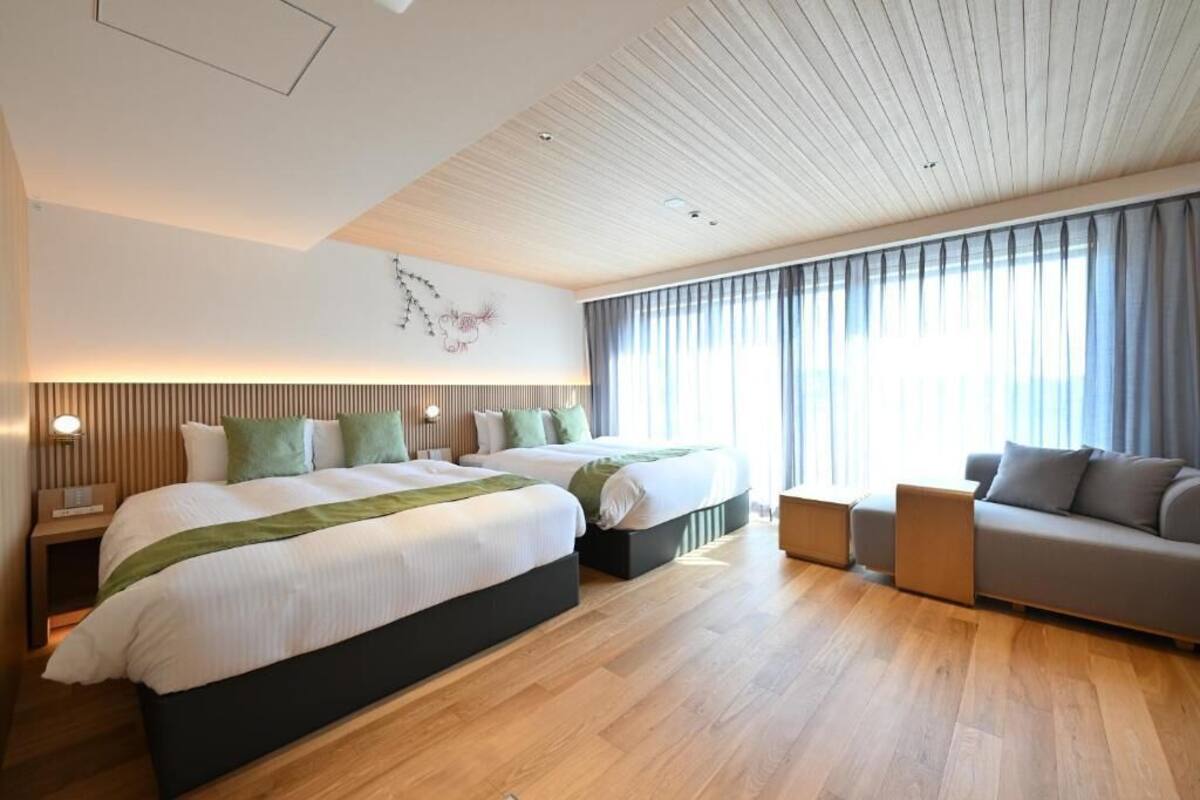 A modern hotel room with two beds, a sofa, wooden flooring, and a large window with curtains.
