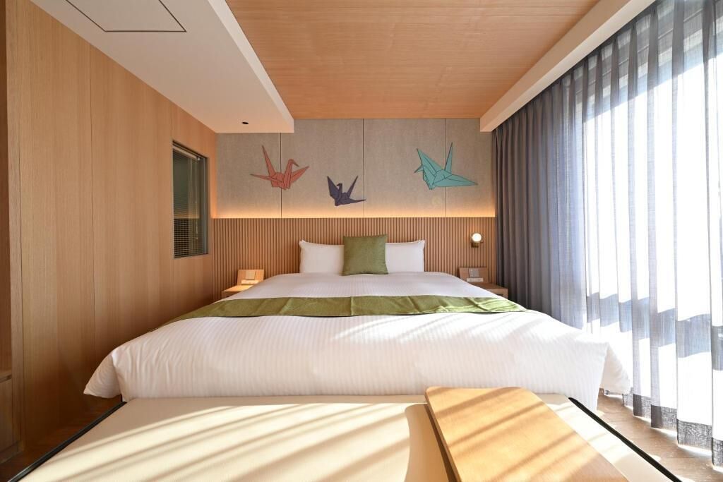 A modern hotel room with a large bed, wooden panel walls, a green throw, and a view of the city through large windows.